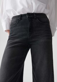 Personne portant un jean noir taille haute et une chemise blanche à boutons rentrées contre un fond uni.