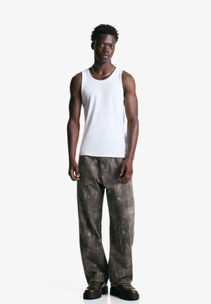 Hvid bomulds tanktop parret med løse, grå strukturerede bukser. Modellen har mørke chunky sko på med grønne snørebånd. Enkel design, neutrale farver.