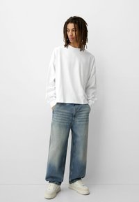 Weißes, langärmliges Sweatshirt mit Rundhalsausschnitt in lockerer Passform, kombiniert mit übergroßen hellblauen Jeans und weißen Sneakers. Einfaches, minimales Design.