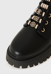Stivale ankle in pelle nera con suola in gomma spessa, caratterizzato da hardware dorato decorato e lacci testurizzati per un ulteriore dettaglio visivo.