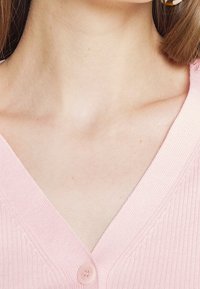 Cárdigan de color rosa claro con un diseño de cuello en V y un cierre de un solo botón, que presenta una textura suave y sutiles detalles de punto.