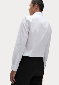 Camisa blanca de manga larga con cuello clásico, textura suave y puños abotonados, llevada con pantalones negros, vista desde la parte de atrás.