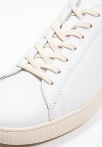 White leather sneaker with round toe, prominent stitching, textured cream-colored sole, and beige laces. Minimalist design with no visible logos.