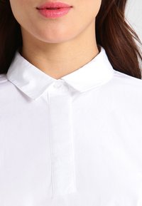 Camicia bianca con colletto, dotata di chiusura a bottoni e tessuto liscio e resistente alle pieghe con una leggera lucentezza. Vista ravvicinata che mette in evidenza il design del colletto.