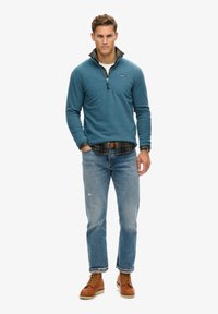 Sudadera de forro polar azul con un cuello con cremallera sobre una camisa de cuadros, combinada con jeans de color azul claro y botas marrones con cordones. Presenta un logo en el pecho.