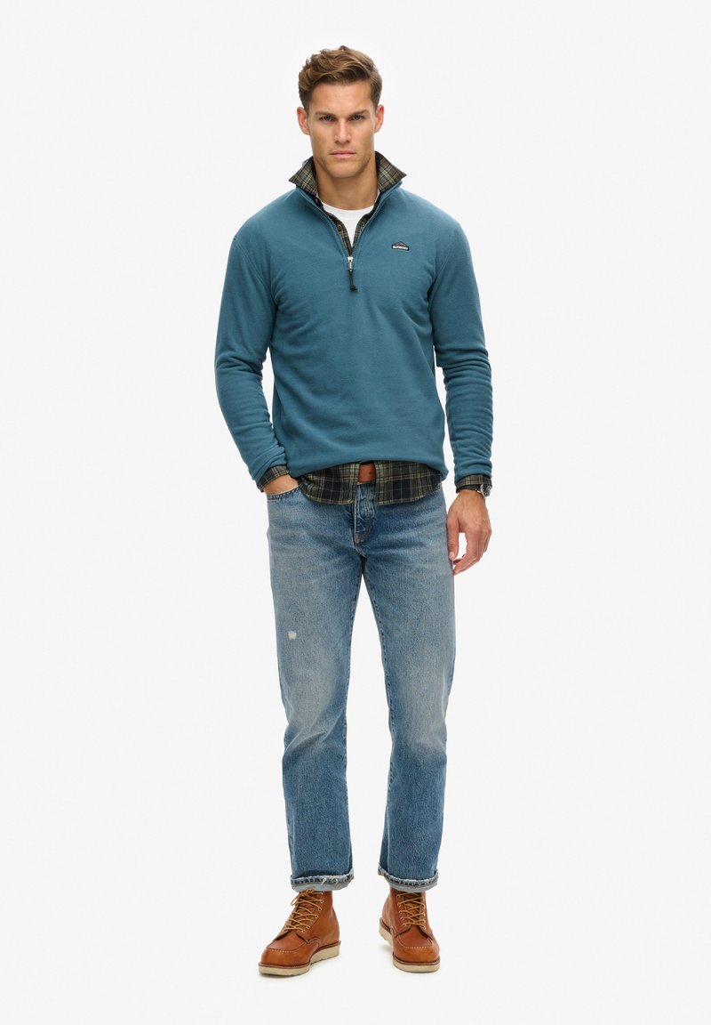 Blauwe fleece trui met een ritskraag over een geruit shirt, gecombineerd met lichtblauwe jeans en bruine veterschoenen. Kenmerkt zich door een logo op de borst.