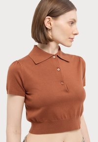 Kortermet, rustfarget polo genser laget av strikket stoff. Har kragehals, tre knapper og en fitted nederkant.