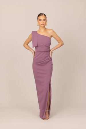 Femme debout, les mains sur les hanches, portant une robe mauve longue jusqu'au sol, asymétrique avec une fente latérale, ainsi que des boucles d'oreilles et un bracelet dorés.