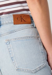 Lichtblauwe denim jeans met een leren Calvin Klein-label op de tailleband. De stof heeft een klassieke denimtextuur met subtiele stikdetails.