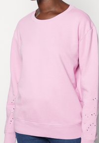 Sudadera de color rosa claro con cuello redondo, mangas largas y detalles de ojal en los puños. Fabricada con suave tela de algodón.