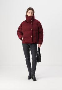 Burgunderfarbene Pufferjacke mit hohem Kragen, goldenen Knöpfen und gesteppter Textur, kombiniert mit schwarzen Jeans und Stiefeletten, während sie eine kleine Tasche hält.