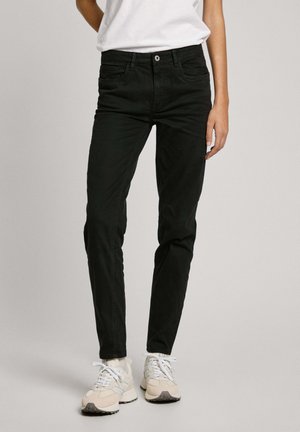 Jeans de denim negro con un ajuste Slim, diseño de cinco bolsillo, costuras visibles y cierre frontal con botón. Combinados con zapatillas de color claro.