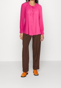 Camicia lunga a maniche rosa brillante con colletto a cravatta, frontale con bottoni e tessuto testurizzato; abbinata a pantaloni larghi marroni e scarpe arancioni.