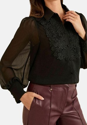 Femme portant un chemisier noir transparent avec un devant en dentelle florale et des manches bouffantes, associé à un pantalon en cuir bordeaux taille haute.