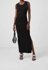Robe maxi noire sans manches avec une fente latérale, associée à des sandales à boucle argentée et un sac à main noir brillant orné de sequins.