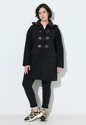 Woman wearing a black coat with toggle buttons, black pants, and black and beige sneakers, standing against a white background.