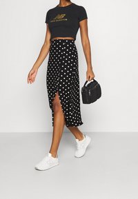 Jupe midi noire à pois avec une fente latérale, associée à un t-shirt court noir avec un logo doré, et des baskets blanches. Petit sac à main noir.