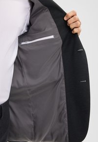 Blazer scuro con una fodera in raso grigia. L'interno presenta una tasca con una cucitura bianca e una texture liscia. All'esterno ci sono dettagli di cucitura sottili.