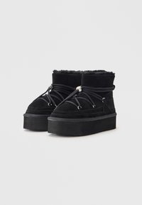 Botas de cano curto em camurça preta com uma sola grossa e plataforma, apresentando atacadores pretos e detalhes em metal prateado. Forro interior suave visível na parte superior.