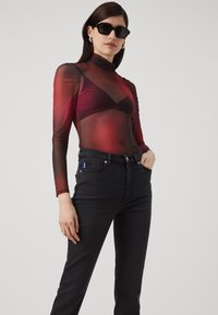 Rød og svart gjennomsiktig langermet topp med tettsittende silhuett, sammen med svarte høye jeans, modelle som har på seg svarte solbriller.