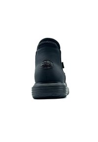 Zapato de cuero negro con forma redondeada, superficie texturizada y tacón prominente. Presenta un detalle de logo y suela de goma con líneas.