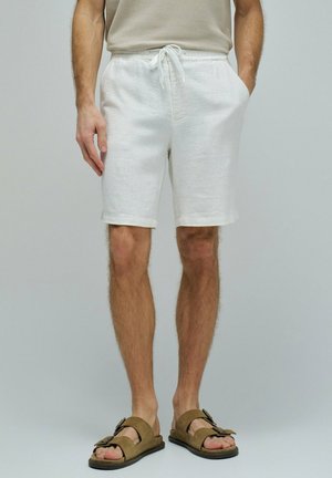 Hombre vistiendo pantalones cortos beige con cordón y bolsillos, sandalias marrones con hebilla y punta abierta, y una camiseta sin mangas beige sobre fondo liso.