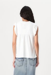 Top in cotone bianco con maniche a volant, caratterizzato da un motivo testurizzato e un collo tondo. Il design sul retro include dettagli decorativi discreti.