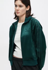 Green velvet Adidas zip-up jacket with embroidered logo, featuring three contrasting stripes along the sleeves, paired with a white top.