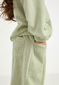 Vihreä fleece-paita, jossa on elastiset hihansuut ja sivutasku. Pehmeä pinta ja rento leikkaus korostavat vapaa-ajan vaatetuksen suunnittelua.