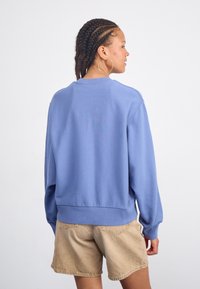 Ljusblå sweatshirt med rund hals, ribbade muddar och nederkant, tillverkad av mjukt tyg, bärs över beige shorts; sedd bakifrån.