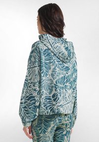 Frau von hinten gesehen, trägt einen Hoodie und eine Hose mit abstrakten Blättermustern in Blau und Beige.