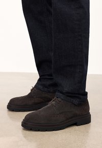 Chaussures en daim marron avec une finition texturée, présentant des détails de brogue, des lacets et une semelle en caoutchouc noir épaisse, assorties à un jean foncé.