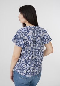 Blusa con stampa floreale in blu navy con fiori bianchi, maniche corte a volant e dettaglio di piega sul retro. Abbinata a jeans azzurri.