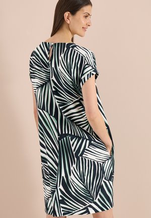 Frau trägt ein kurzärmeliges schwarz-weißes Kleid mit Blattmuster, steht seitlich mit den Händen in den Taschen vor einem beigen Hintergrund.