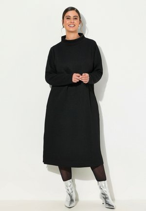 Schwarzes langärmeliges Kleid mit hohem Kragen und strukturiertem Muster, kombiniert mit glänzenden silbernen Stiefeletten und schwarzen Leggings.