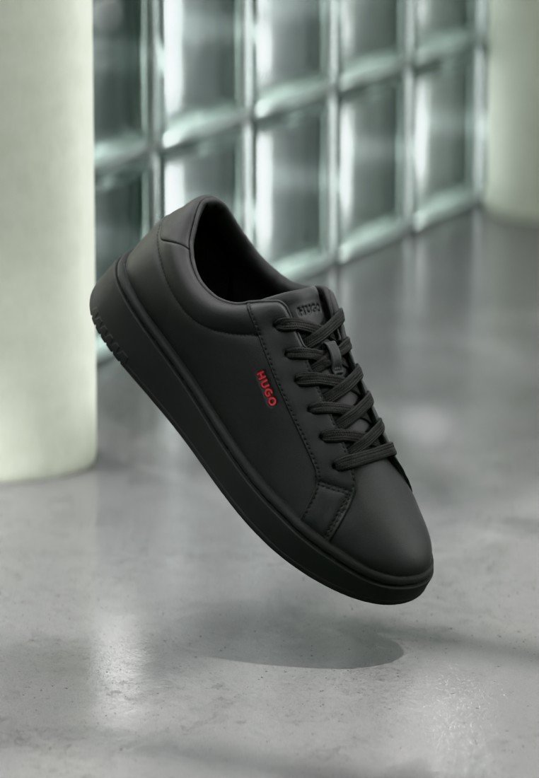 Baskets basses noires avec lacets noirs et logo rouge "HUGO" sur le côté, placées devant un fond de blocs de verre réfléchissants.