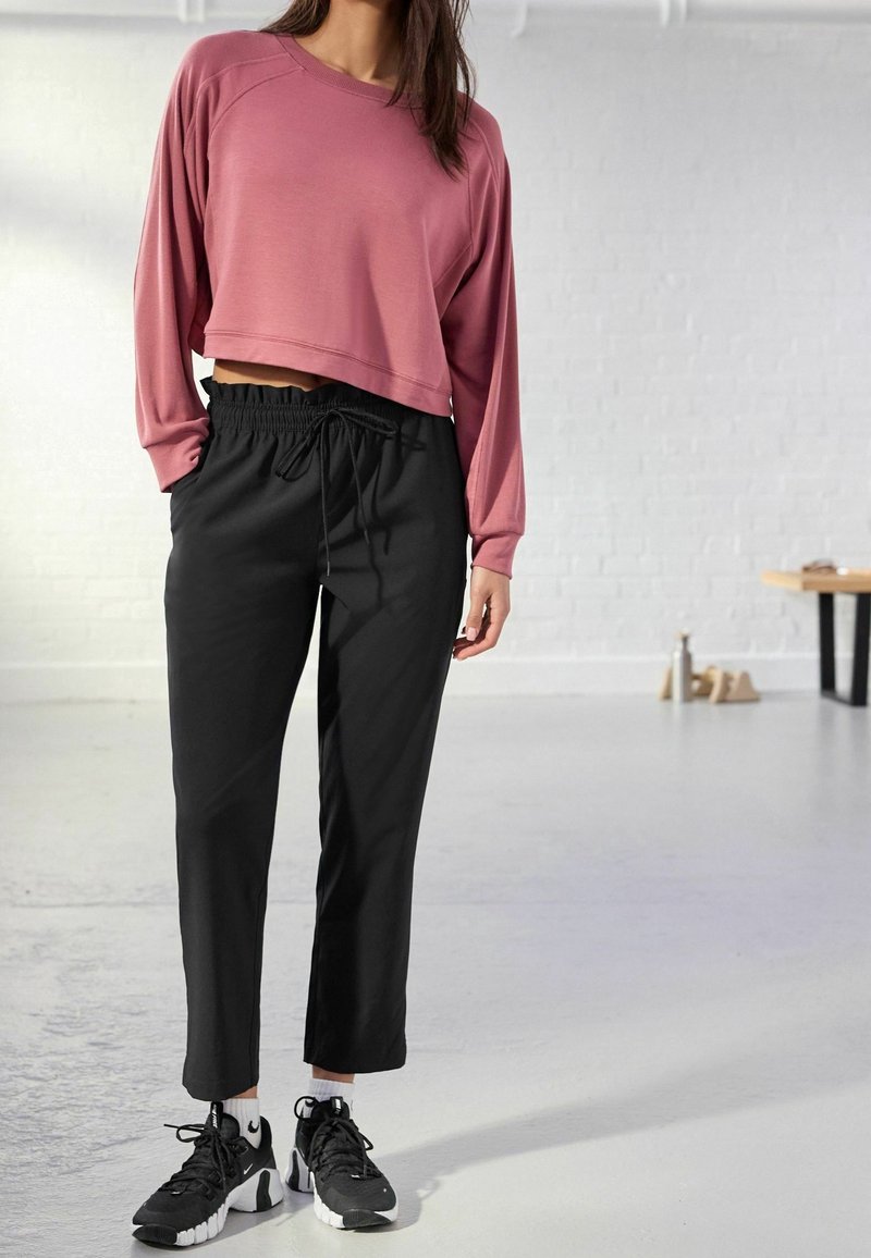 Sweat-shirt court rose à manches longues porté avec un pantalon noir à cordon de serrage. Le mannequin porte des baskets noires avec des accents blancs.