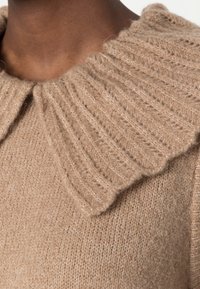 Primo piano di una persona che indossa un maglione lavorato a maglia beige con un ampio colletto a coste ondulato.