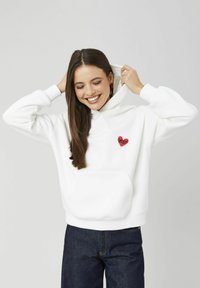 Sweat à capuche blanc avec une poche avant et un écusson en forme de cœur rouge. Texture lisse, coupe décontractée. Associé à un jean bleu foncé.