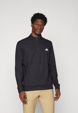 Pull noir à manches longues avec fermeture à demi-éclair et logo Adidas blanc sur la poitrine. Texture lisse avec un design ajusté.