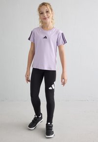 Fille se tient contre un mur blanc portant un t-shirt de sport violet clair, un legging noir avec un logo blanc et des baskets noires avec des semelles blanches.