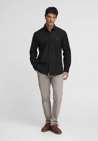 Zwarte overhemd met lange mouwen en een button-down kraag, voorzien van knoopsluiting aan de voorkant en een klein logo op de borst, gecombineerd met lichtgekleurde broeken en bruine schoenen.