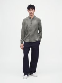 Un homme debout portant une chemise grise à manches longues à motifs, un pantalon ample foncé et des baskets blanches sur un fond clair uni.