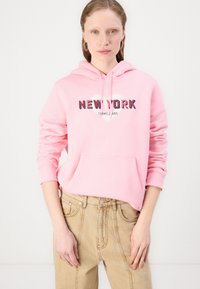 Felpa leggera rosa con la grafica "NEW YORK" e "TOMMY JEANS" in bianco e nero, dotata di tasca frontale e maniche lunghe. Abbinata a pantaloni beige.