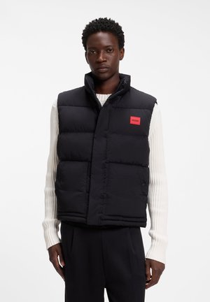 Mørkhudet person iført en sort polstret vest med rødt Hugo-logo over en hvid ribstrikket sweater, stående foran en ensfarvet hvid baggrund.
