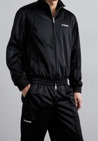 Black tracksuit made of smooth, lightweight fabric. Features a high collar, zip closure, and elastic cuffs and waistband. White logo detail.