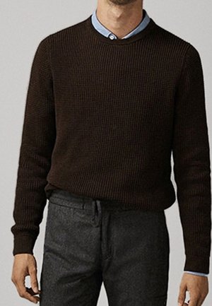 Homme portant un pull en maille brun foncé par-dessus une chemise à col bleu clair et un pantalon gris foncé, debout devant un fond neutre.