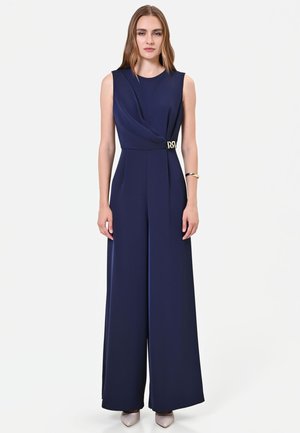 Donna in piedi che indossa una tuta senza maniche blu navy con gambe larghe e un piccolo dettaglio con fibbia dorata in vita, abbinata a scarpe chiare.