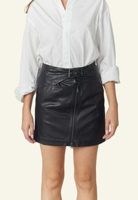 Zwarte leren mini rok met een ritssluiting aan de voorkant, een ceintuurlijn en details aan de zijkant. Gecombineerd met een witte blouse met knopen.