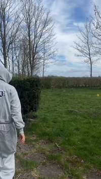 Άτομο με γκρι φούτερ με κουκούλα και παντελόνι περπατάει σε μονοπάτι με χορτάρι κοντά σε κλαδεμένους φράχτες και δέντρα χωρίς φύλλα κάτω από ένα συννεφιασμένο ουρανό.